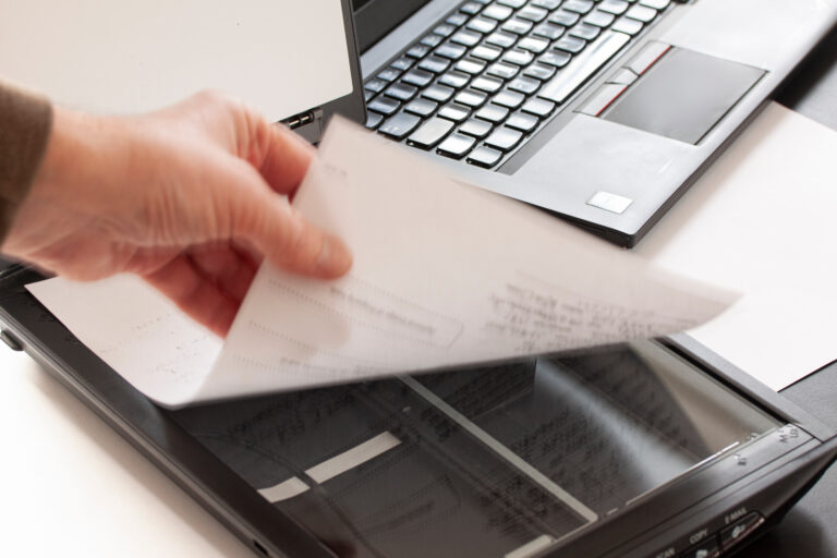 A hand puts a piece of paper on a flatbed scanner with a laptop computer in the background. Selective focus and hand in motion blur