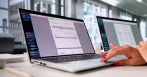 Two open laptops on a white desk showing invoice documents on-screen as a person types.